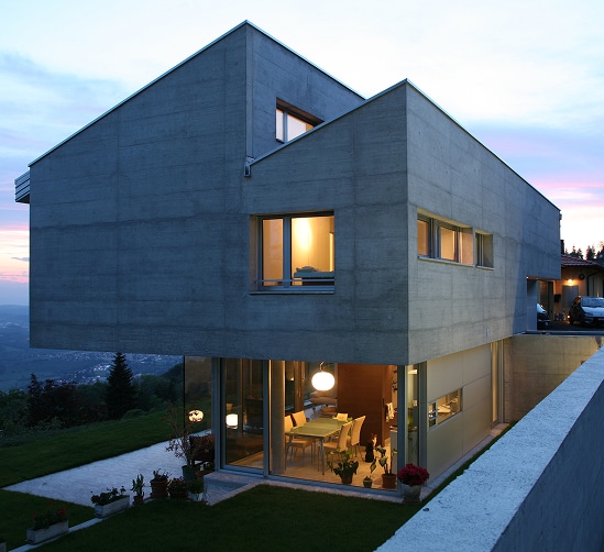 Architecture contemporaine en béton au crépuscule, surplombant une colline. Intérieur chaleureux illuminant la salle à manger vitrée.