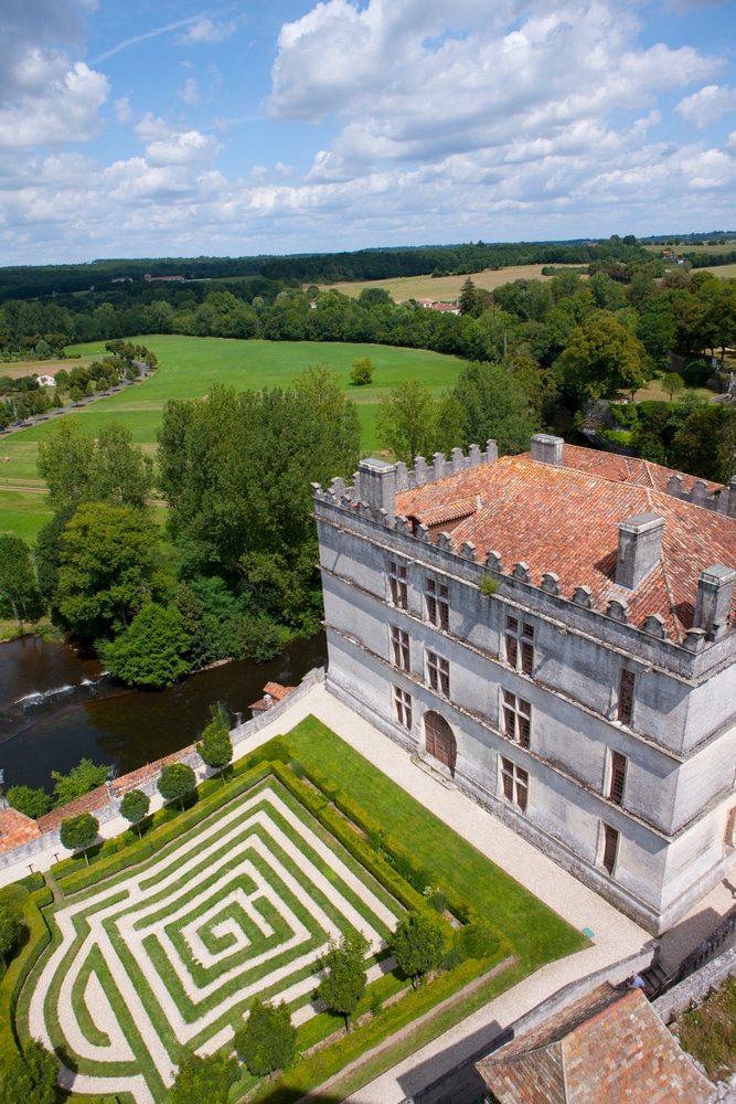 Vue plongeante sur un château ancien en pierre avec créneaux et un toit de tuiles. Un jardin labyrinthe spiralé borde la façade. Nature luxuriante et rivière.