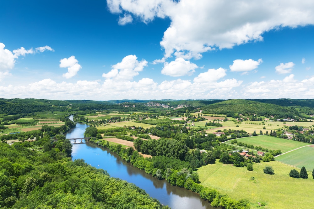 Rivière, vallée verdoyante et ciel bleu : paysage de France Vue aérienne d'une vallée verdoyante : rivière sinueuse, pont, champs agricoles et maisons rurales sous un ciel d'été très bleu et nuageux.