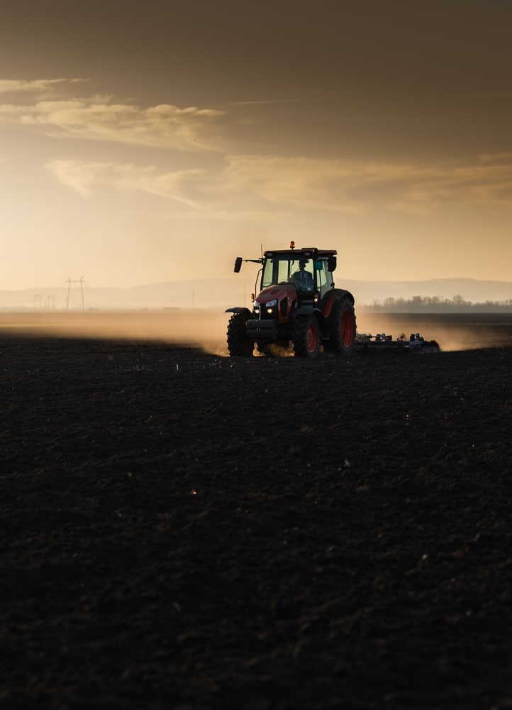 Tracteur en action : Labourage du champ au coucher du soleil Tracteur rouge laboure un champ sombre au coucher du soleil, soulevant de la poussière dorée. Lignes électriques à l'horizon.