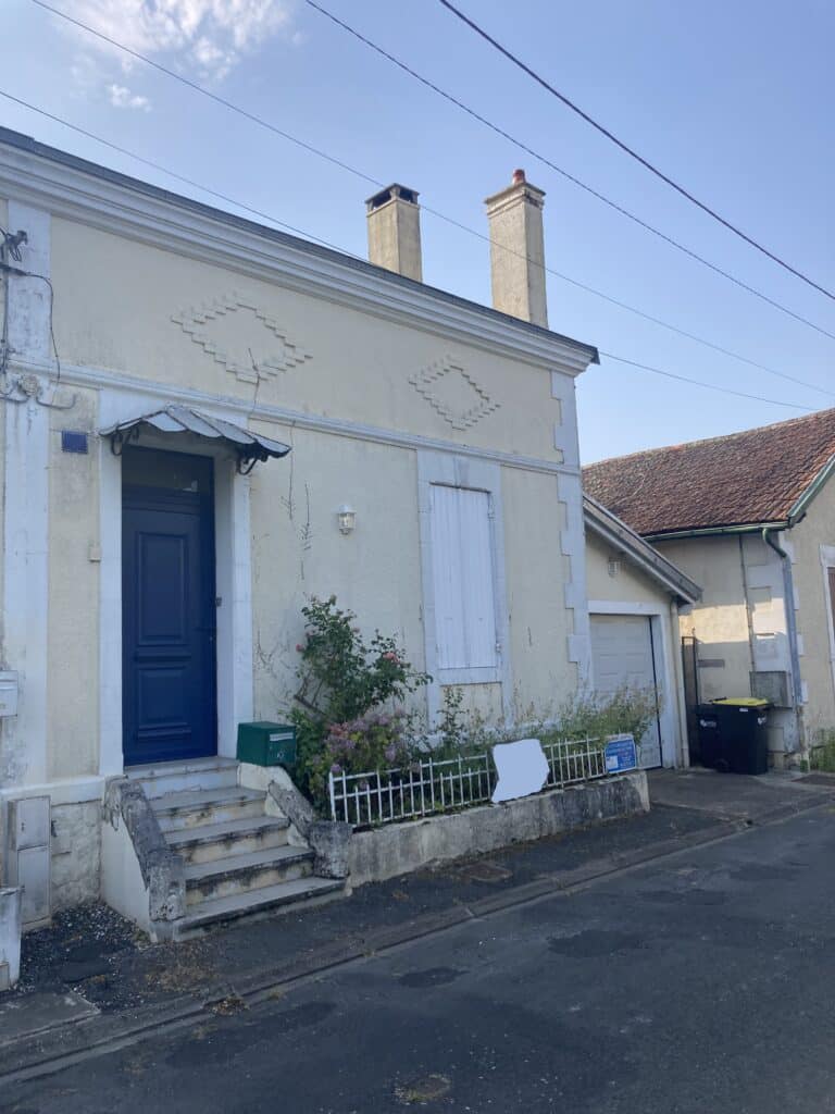Façade de maison crème avec porte bleue, volets blancs et jardinet fleuri sous un ciel bleu.